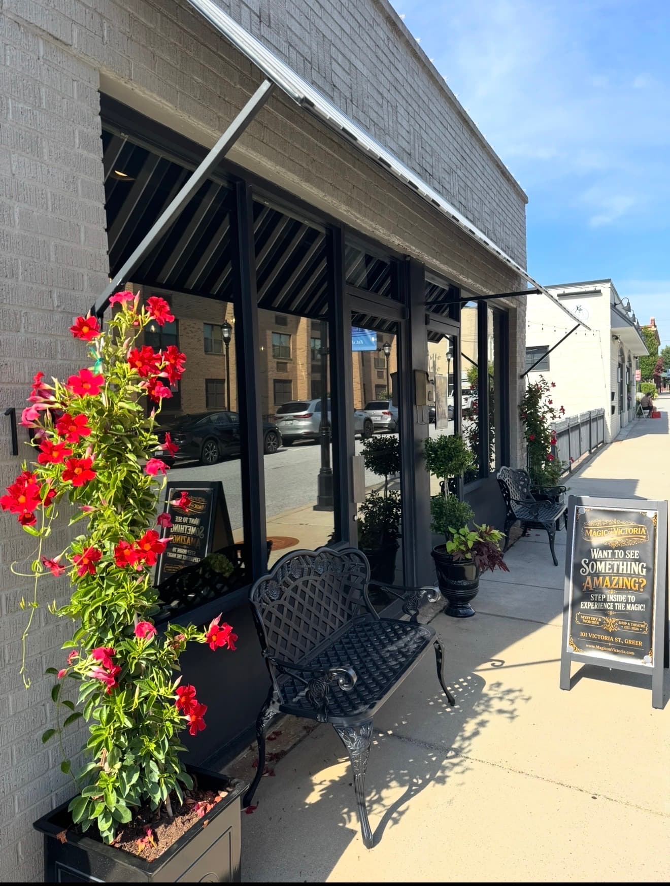 The Magic on Victoria storefront by day on Victoria Street, a flower-lined bench beneath the awning and a sidewalk sign reading Want to See Something Amazing at 101 Victoria Street, Greer